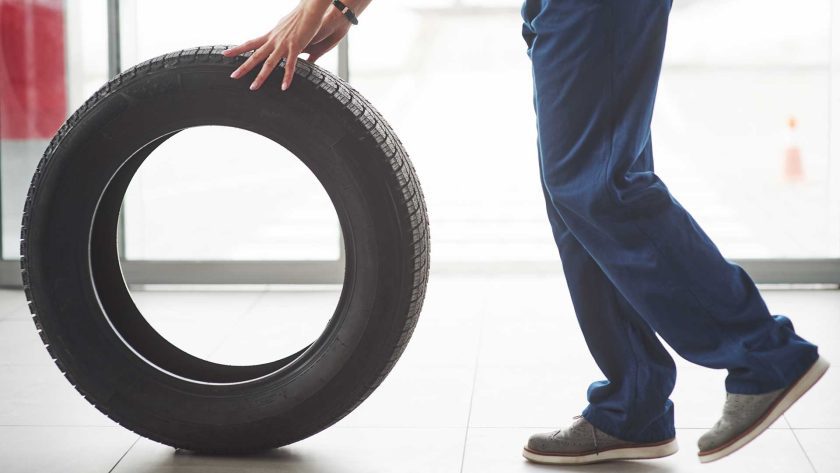Technician rolling a black replacement tire across the tiled floor of a tire and automotive mechanic shop, showing the tire’s tread pattern and the mechanic’s blue work pants and gray shoes.