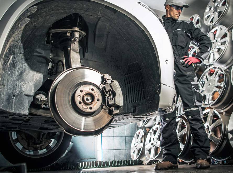 Auto technician standing beside a lifted car with the wheel removed, inspecting the brake rotor and suspension system in a tire and wheel service shop.